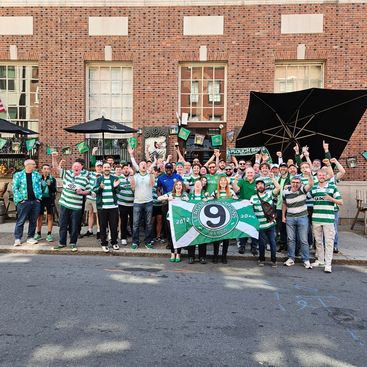 Plough Bhoys Celtic Supporters Club members in green-and-white hoops holding their banner outside The Plough and The Stars in Philadelphia