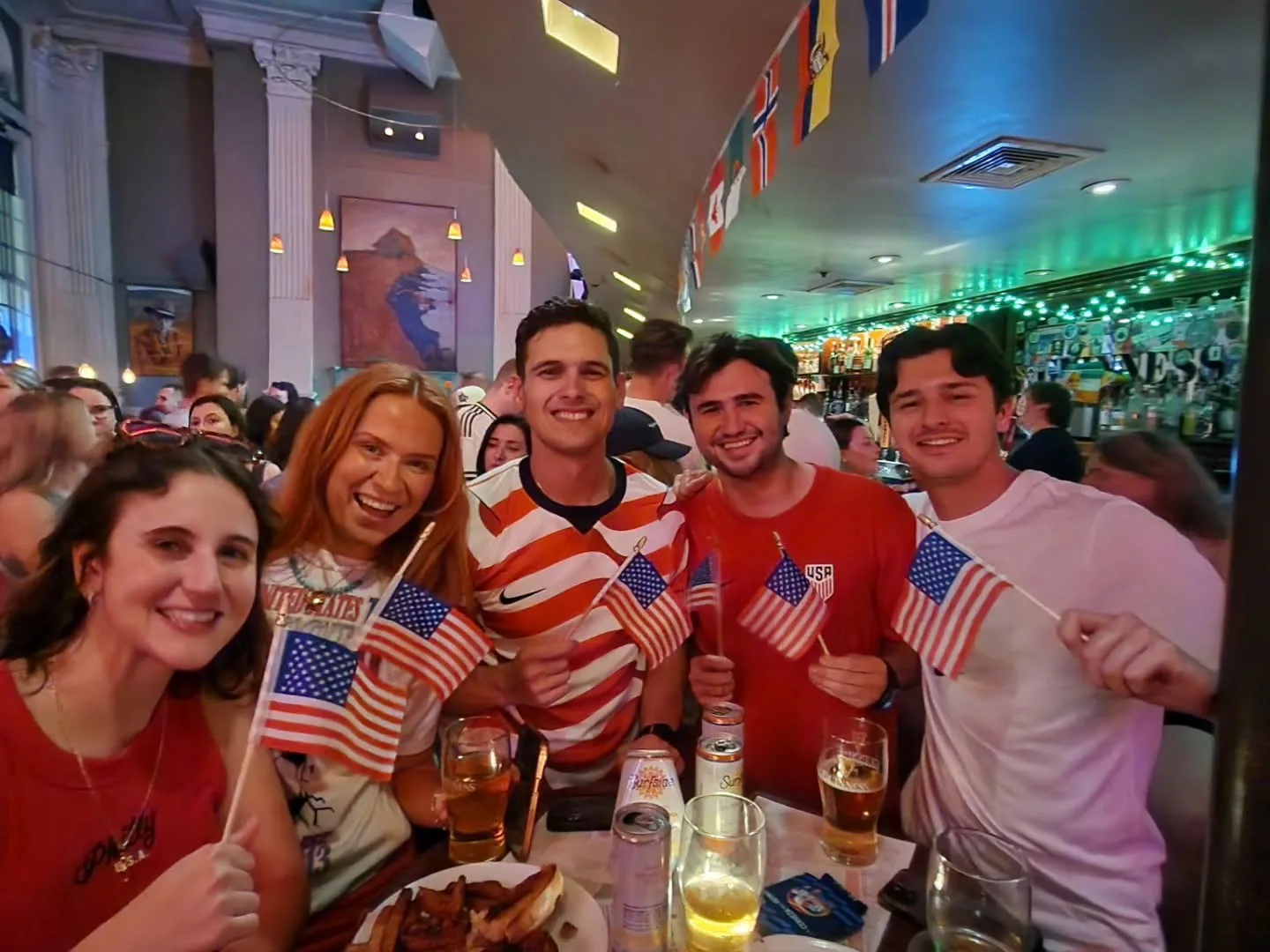 USA soccer fans in jerseys waving American flags and enjoying beers at The Plough and The Stars during a World Cup match, international flags strung overhead